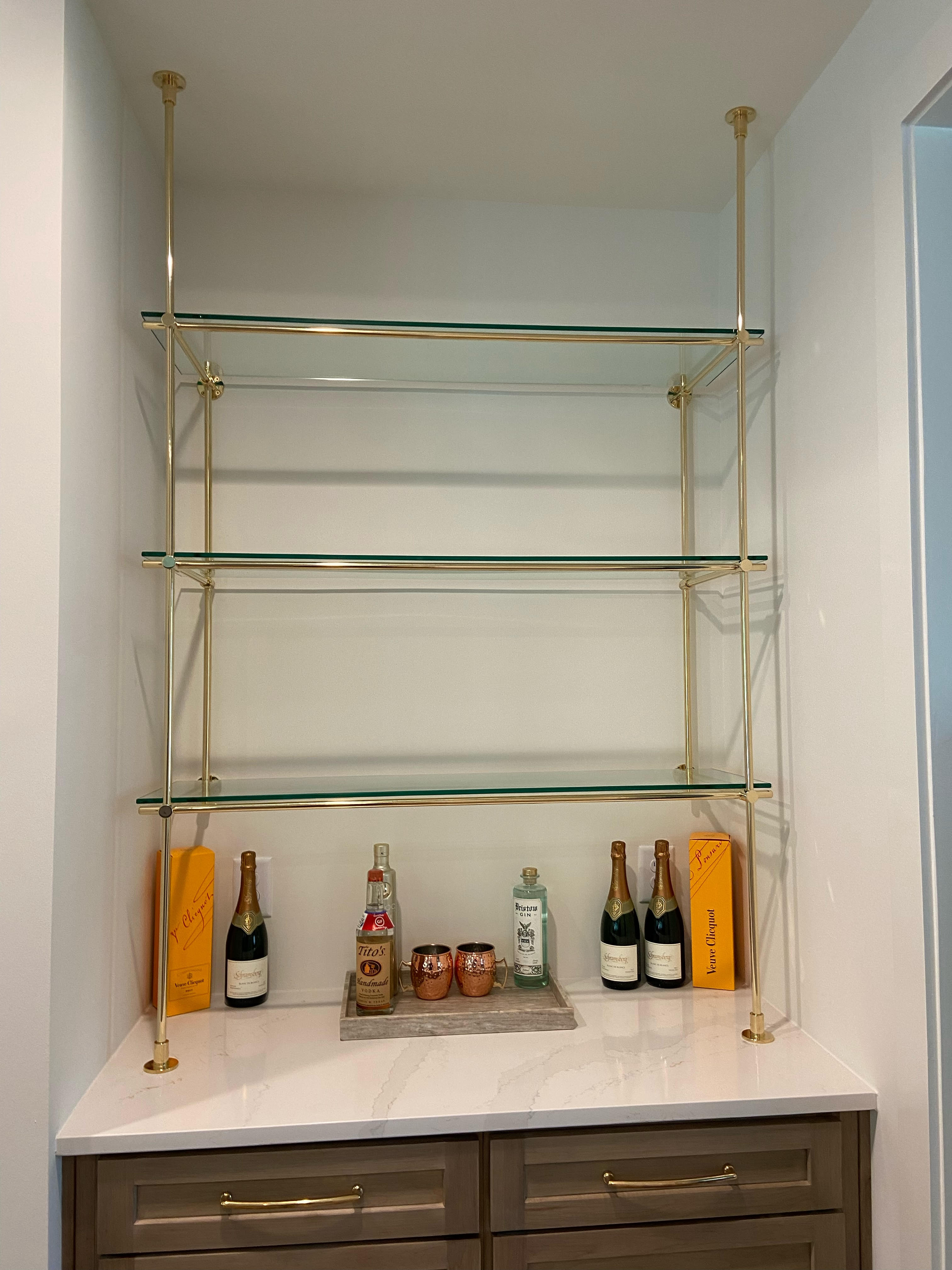 A luxurious countertop-to-ceiling mount shelving unit made of solid brass, featuring three glass shelves. The unit is elegantly displayed in a home bar area, adorned with premium spirits and champagne bottles, reflecting a sophisticated and modern design. Perfect for showcasing collectibles or creating a stylish storage solution.