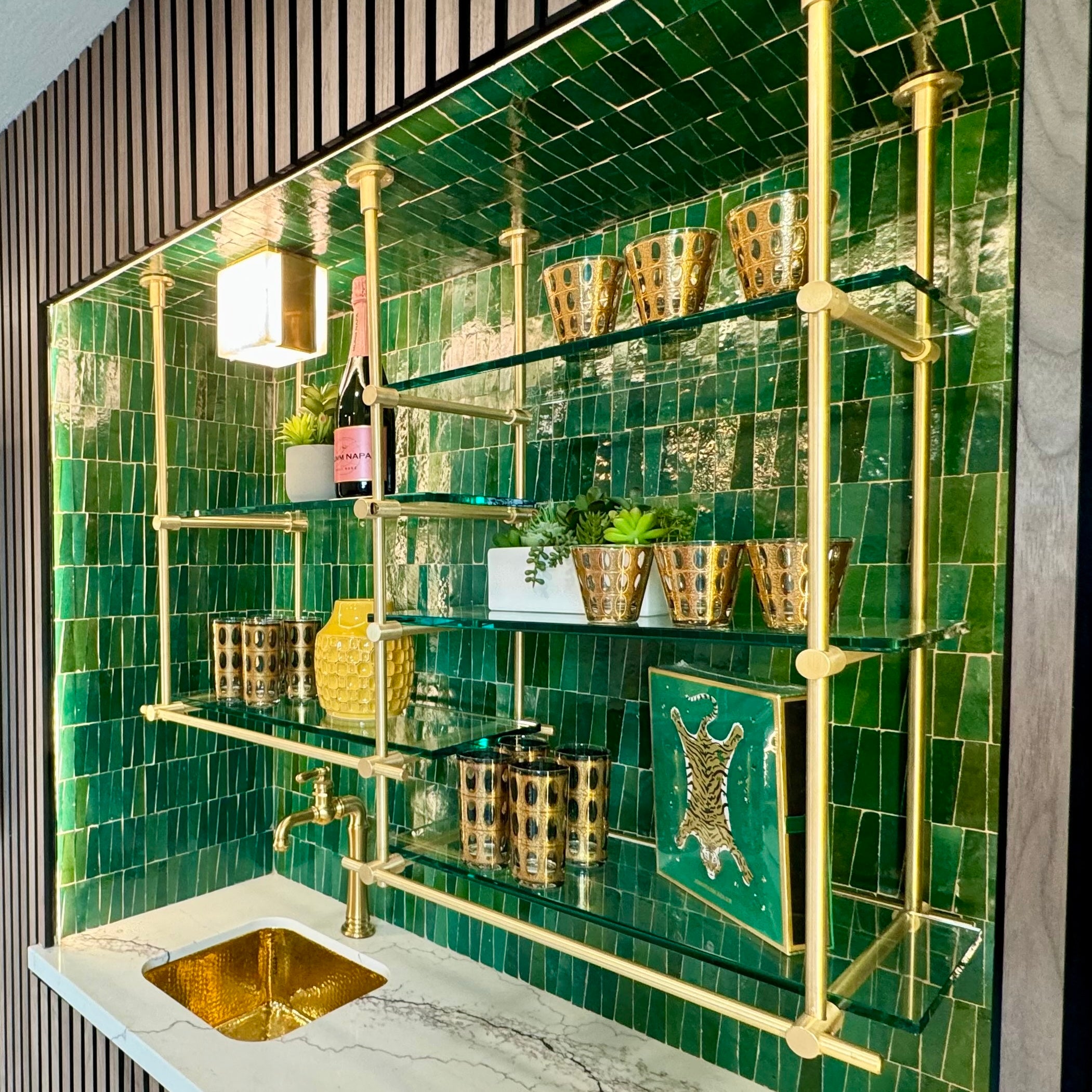 Luxurious brass shelving unit with glass shelves, set against a striking green tiled backsplash. This sophisticated shelving system is perfect for displaying decorative items and drinkware. Features include a built-in brass faucet and sink, enhancing the elegant and functional design of modern kitchens and bars.